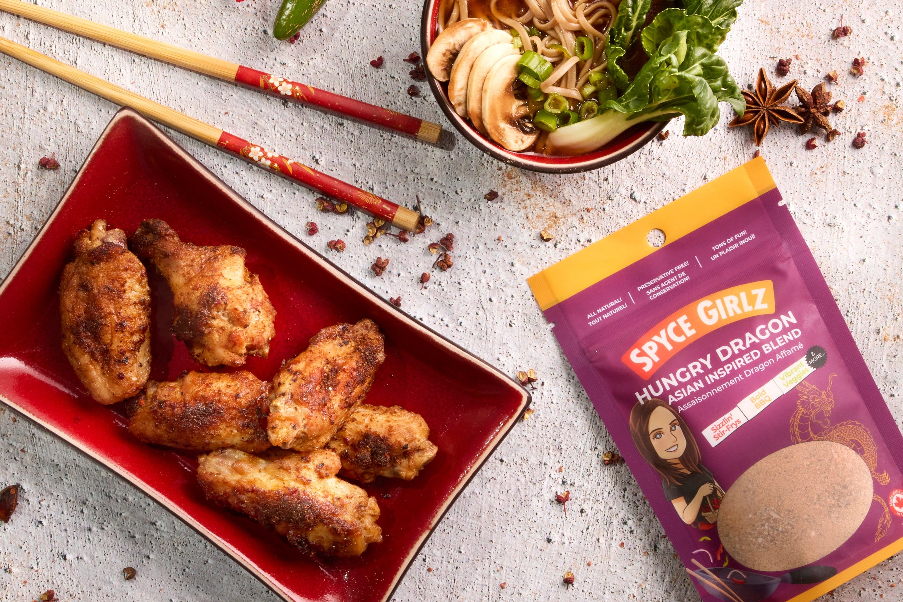 Asian chicken wings on a red plate with a package of 'Spyce Girlz' seasoning mix, surrounded by spices and a bowl of noodles.